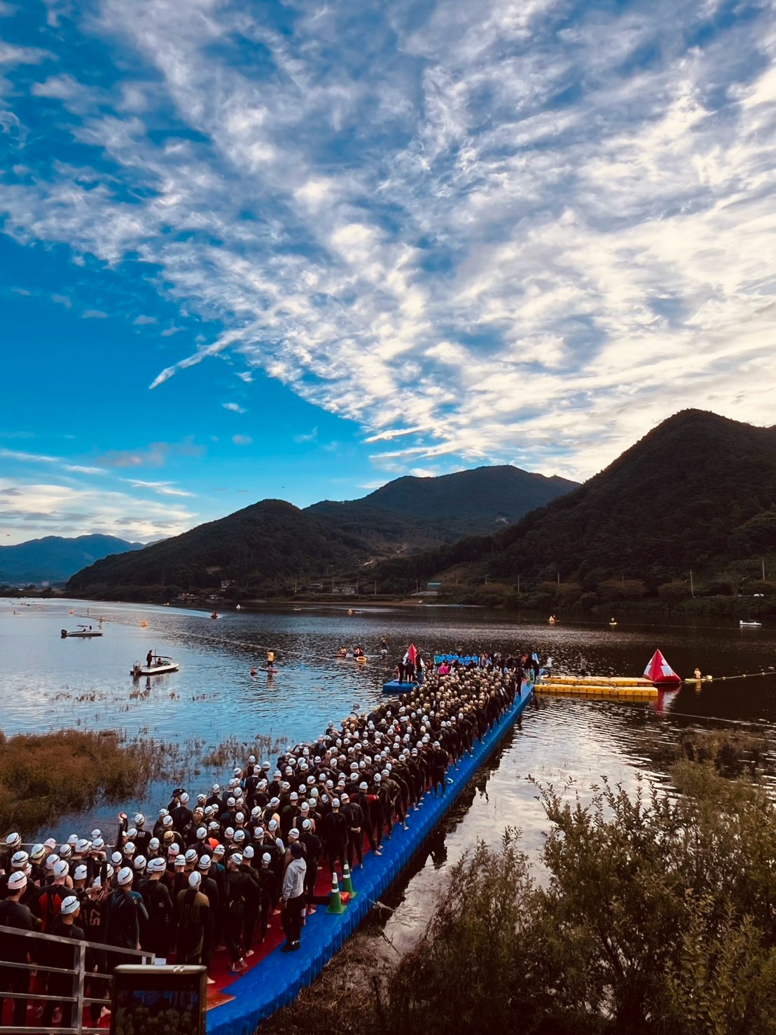 戴泳帽的選手在藍色浮橋排隊，後方是湖面、山景與雲層